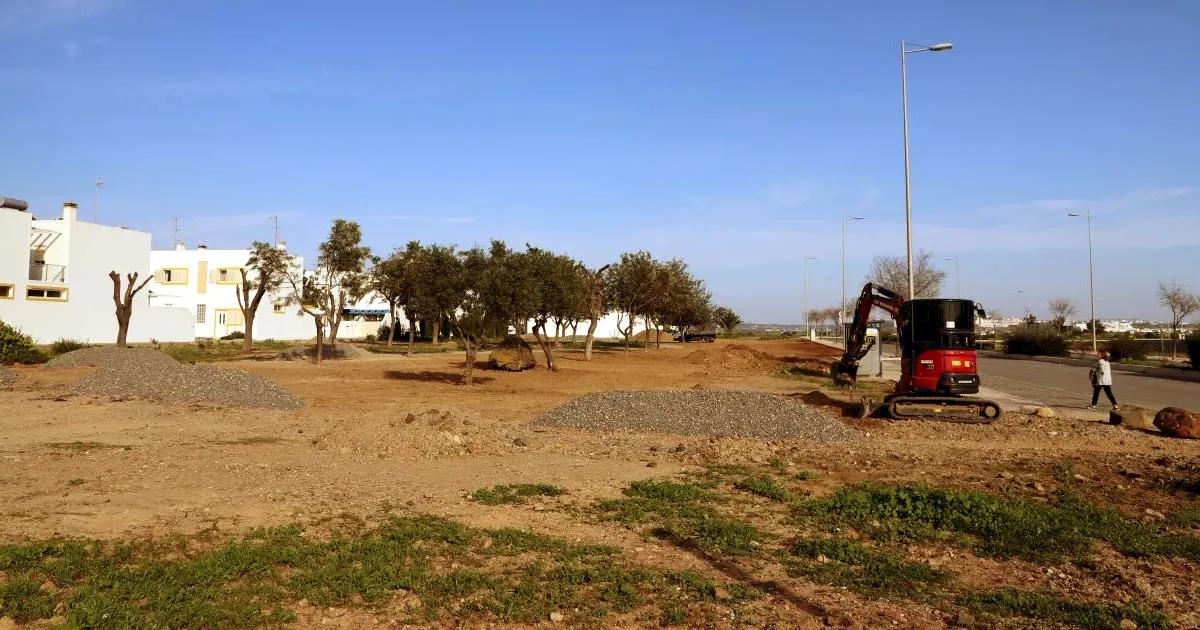 Obras urbanas com escavadora e casas ao fundo.