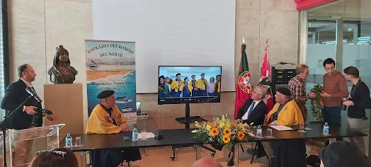 Cerimónia oficial em sala com várias pessoas e bandeiras.